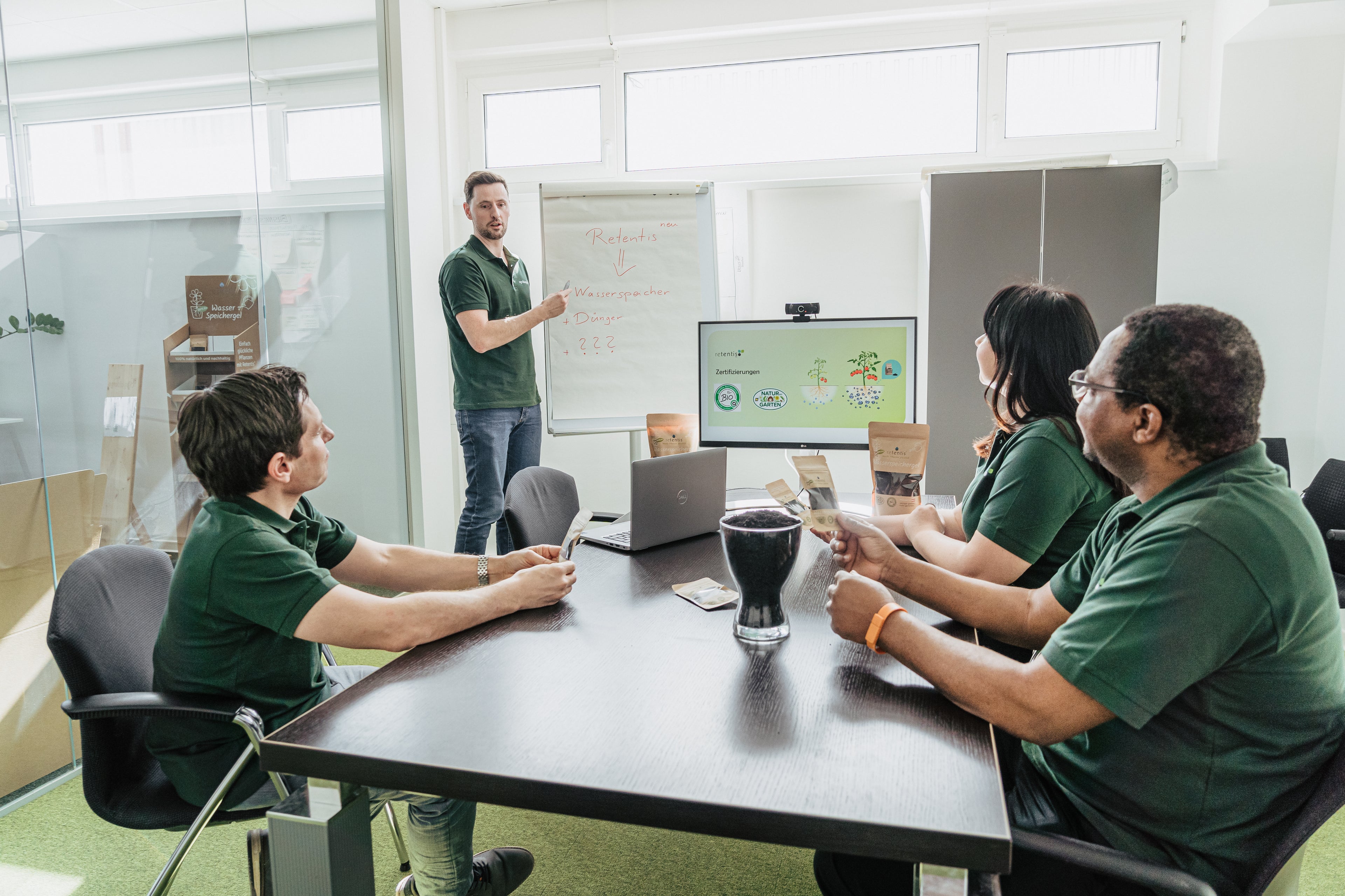 Team von Agrobiogel bei einer Besprechung: Vier Personen sitzen am Konferenztisch, während ein Kollege am Flipchart präsentiert, unterstützt durch eine Bildschirmpräsentation mit Produkt- und Nachhaltigkeitssymbolen.