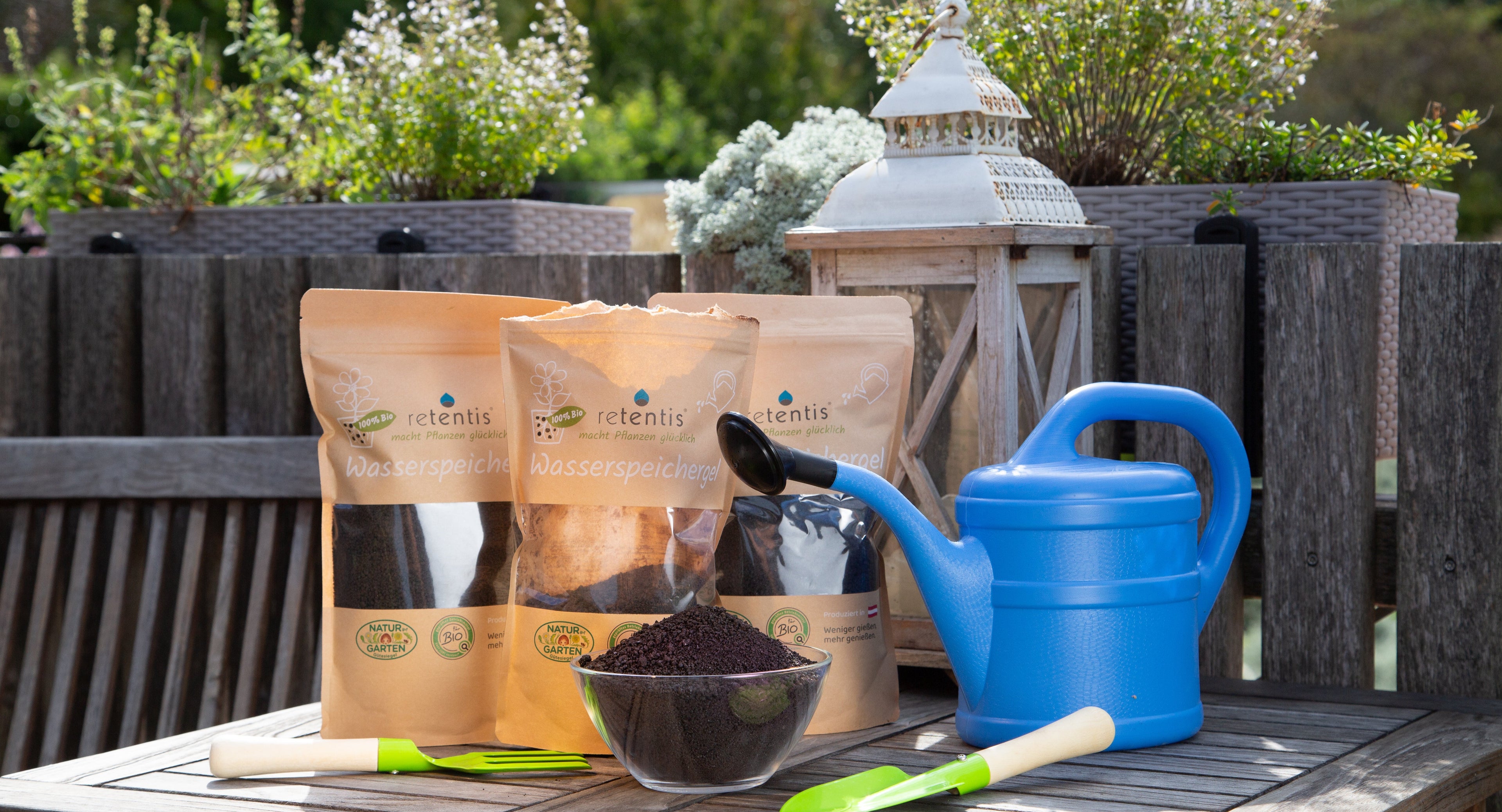 RETENTIS® Agrobiogel Wasserspeichergel für nachhaltige Gartenpflege: Bio-Granulat zur Bodenverbesserung und Wasserspeicherung, mit Verpackungen, Schale, Gartengeräten und Gießkanne auf Holztisch im Garten.