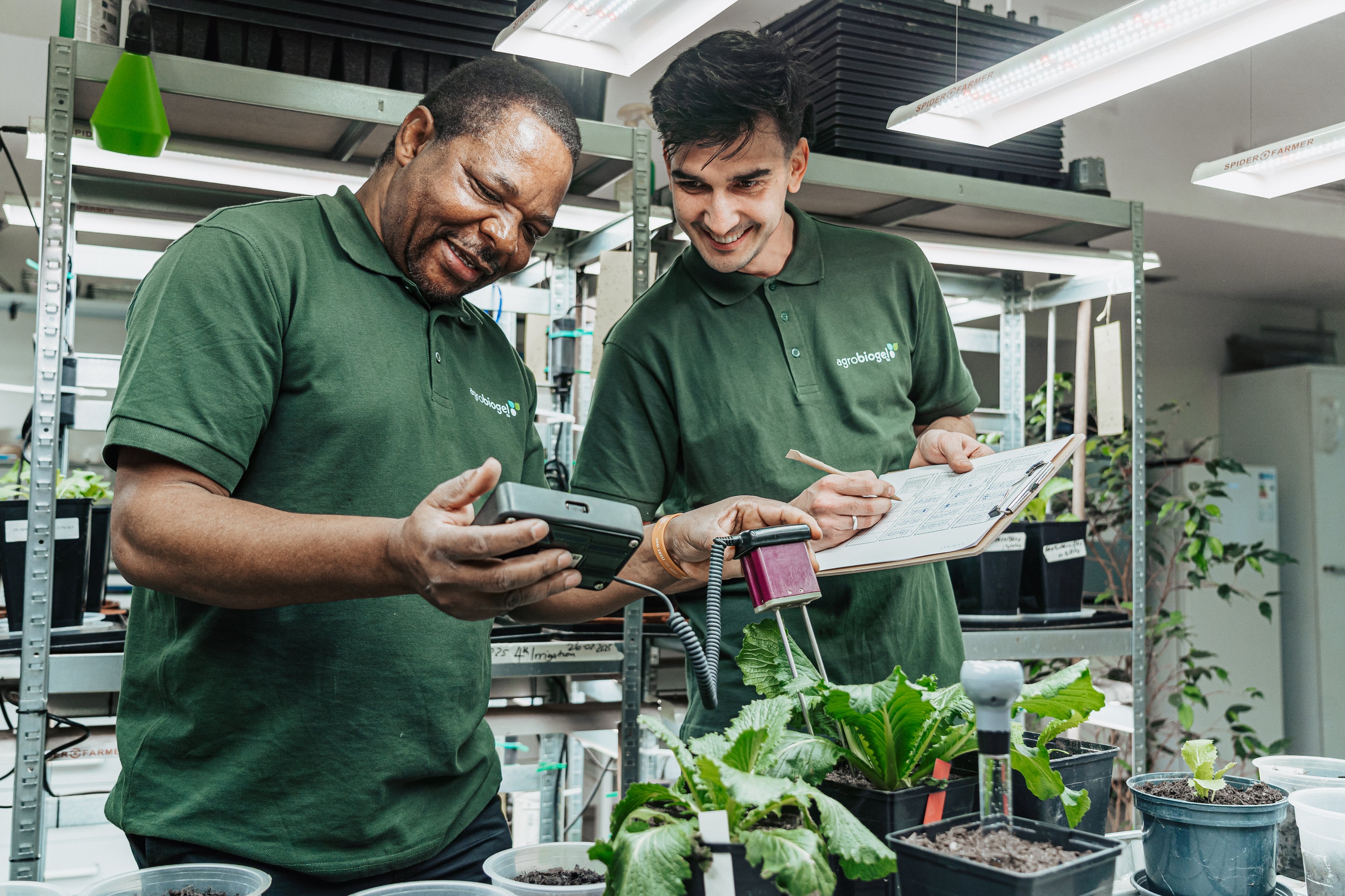 Zwei Mitarbeiter von Agrobiogel arbeiten in einer Indoor-Pflanzenanzuchtanlage, messen Werte mit einem Handgerät und notieren Ergebnisse auf einem Klemmbrett, während sie gesunde grüne Pflanzen begutachten.